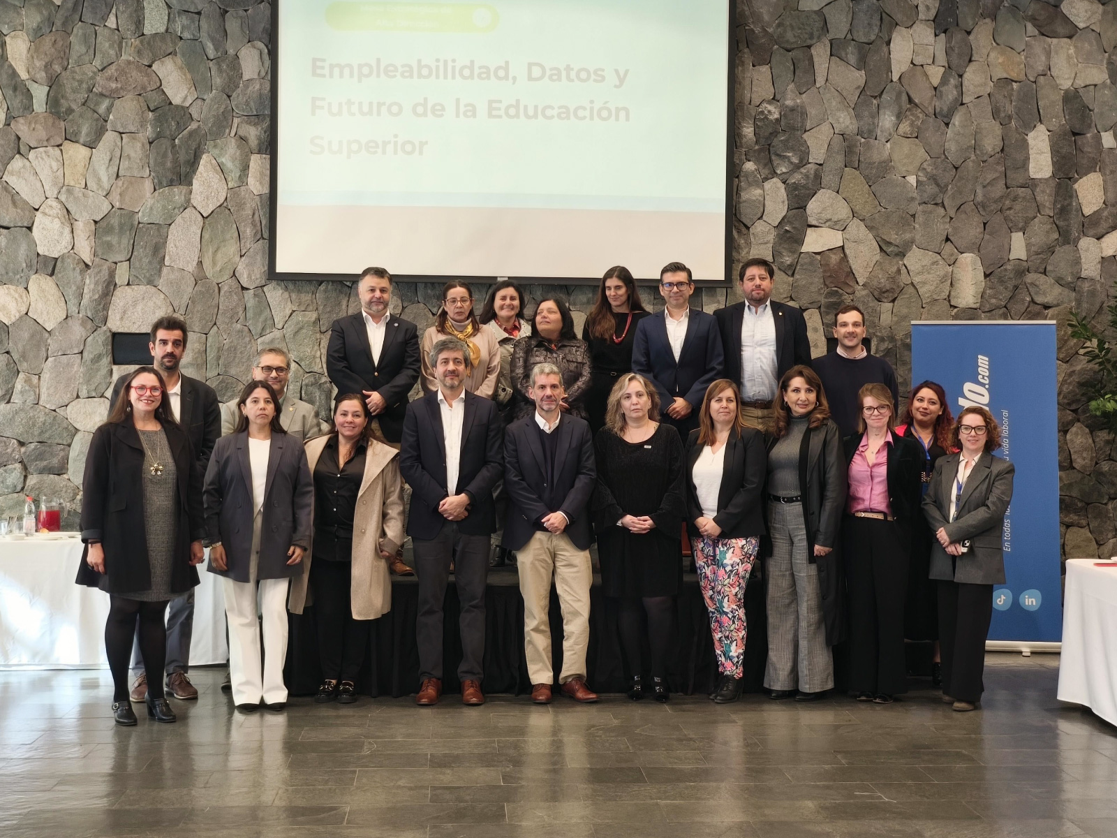 About 20 professionals in business attire stand in two rows for a group photo in a conference space with a stone wall backdrop and a projection screen displaying a Spanish title.