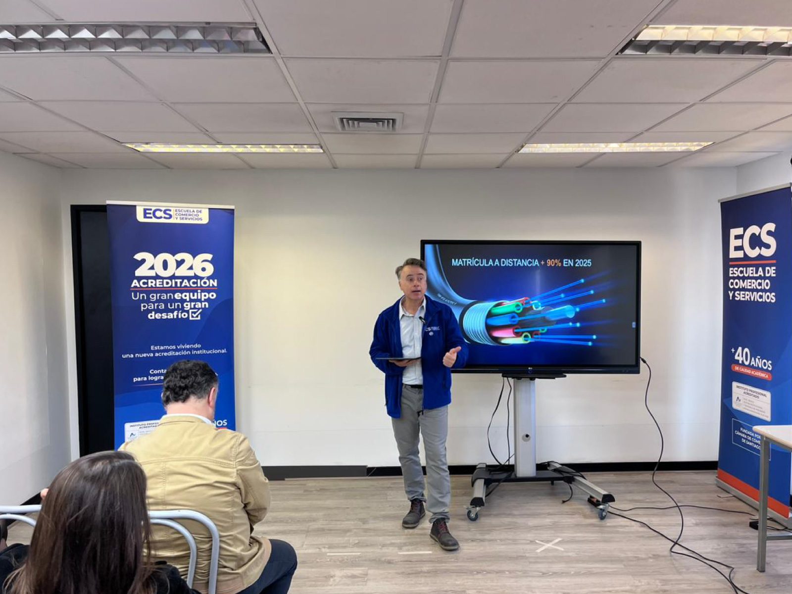 Lecturer in a blue jacket presenting in a classroom with a large screen showing fiber-optic graphics; banners on both sides read ECS and 2026 Acreditación.