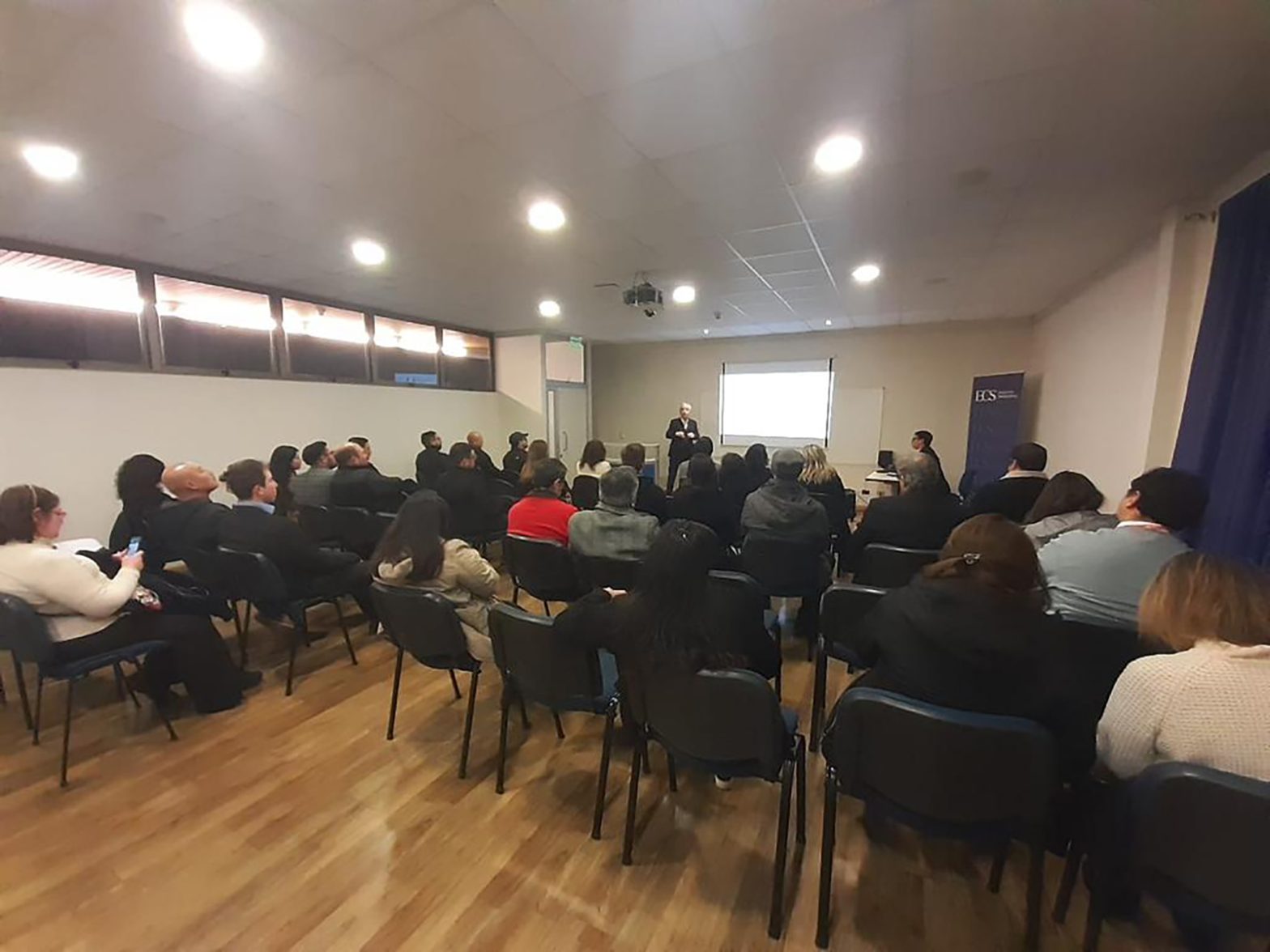 Conference room with rows of chairs and attendees watching a speaker at the front; projector screen behind him.