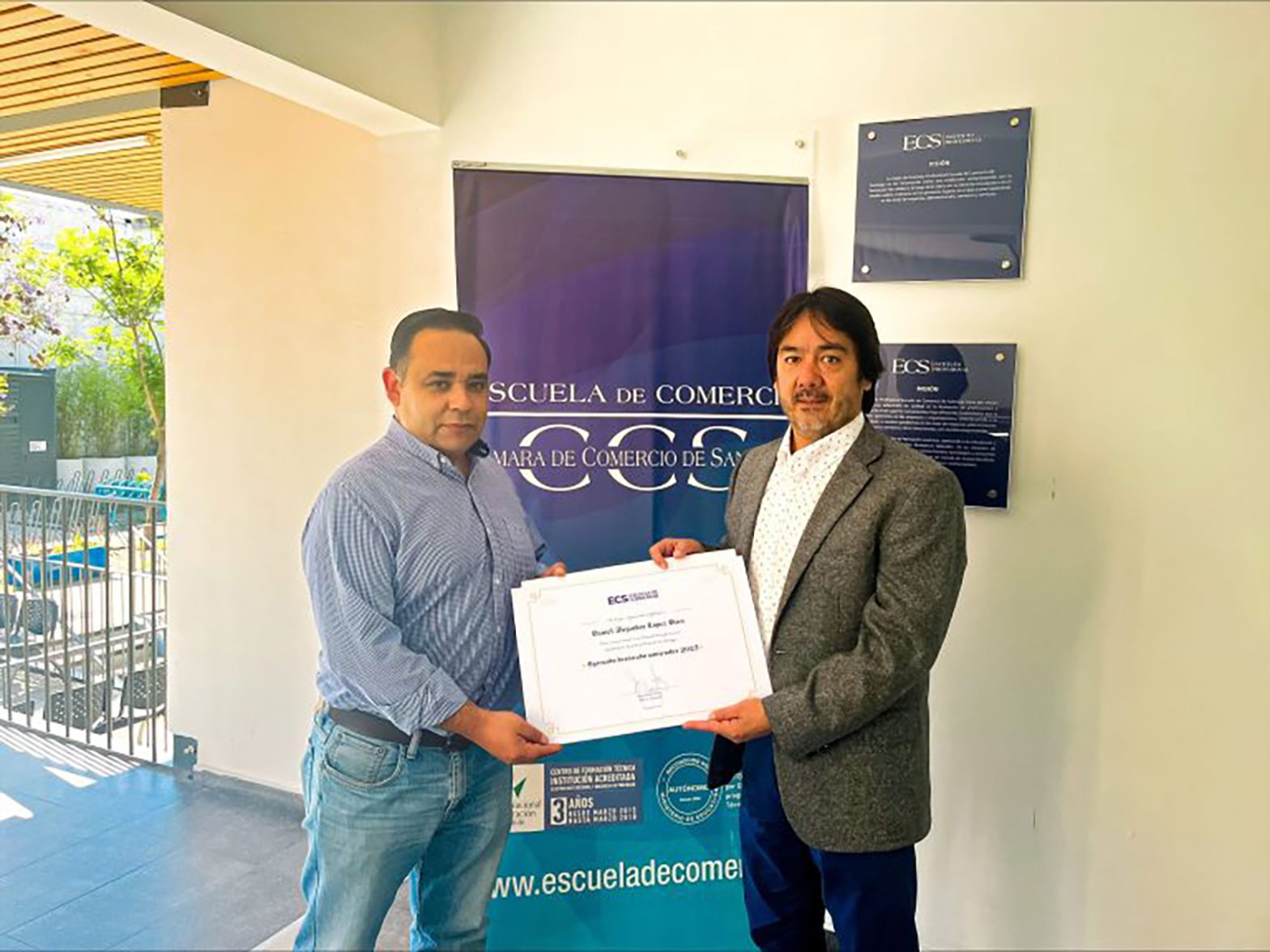 Two men pose indoors holding a certificate in front of a blue Escuela de Comercio banner on the wall behind them.