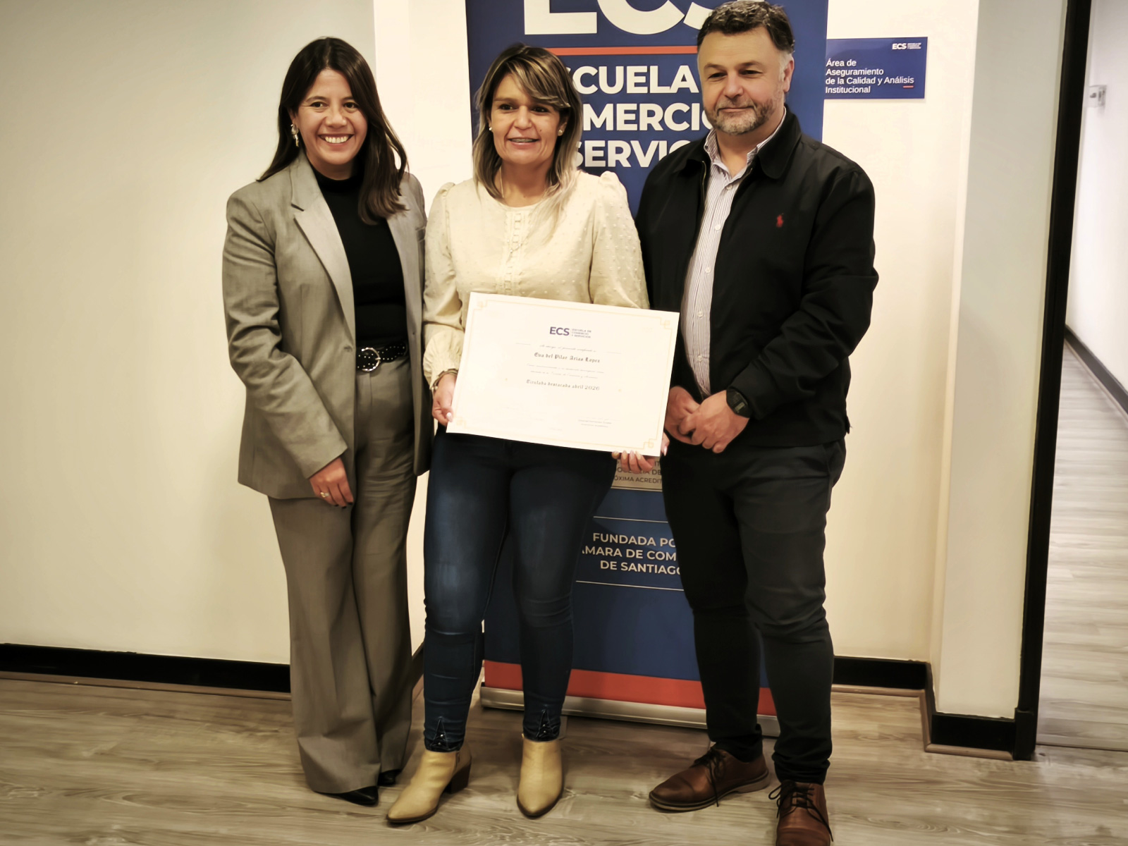 Three adults stand indoors and pose with a certificate, smiling in front of a blue banner.