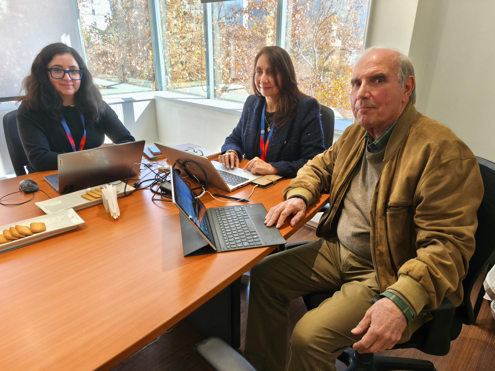 Three colleagues sit around a wooden conference table with laptops, papers, and snacks during a daytime meeting in a bright office windowed room with autumn trees outside.