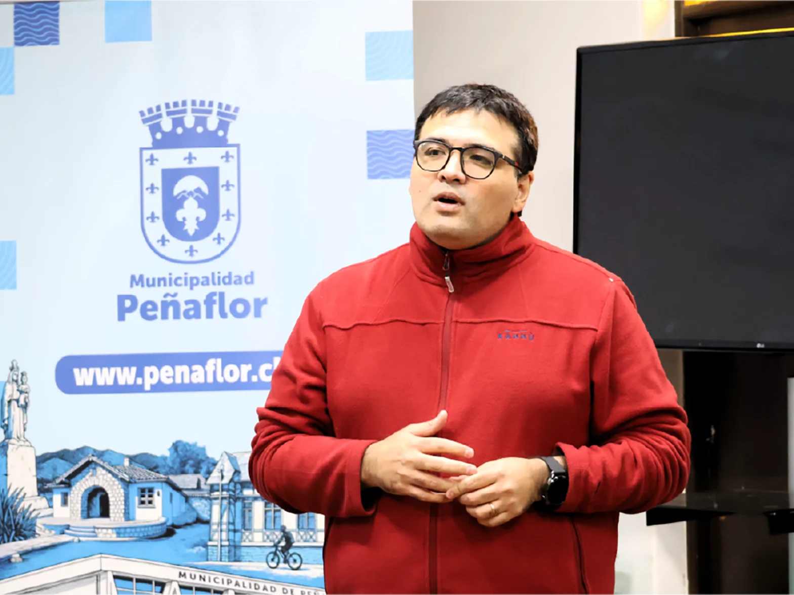 Man in a red jacket giving a talk in front of a Peñaflor municipality banner under a crest logo