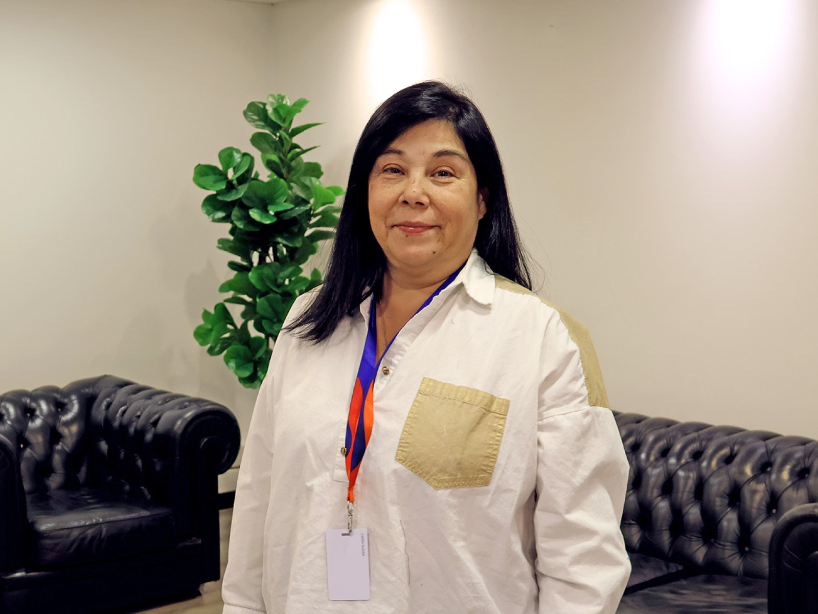 A smiling woman with shoulder‑length dark hair stands in a lobby or lounge area, wearing a white shirt with a beige pocket and a lanyard with an ID badge, behind black leather couches and a green plant.