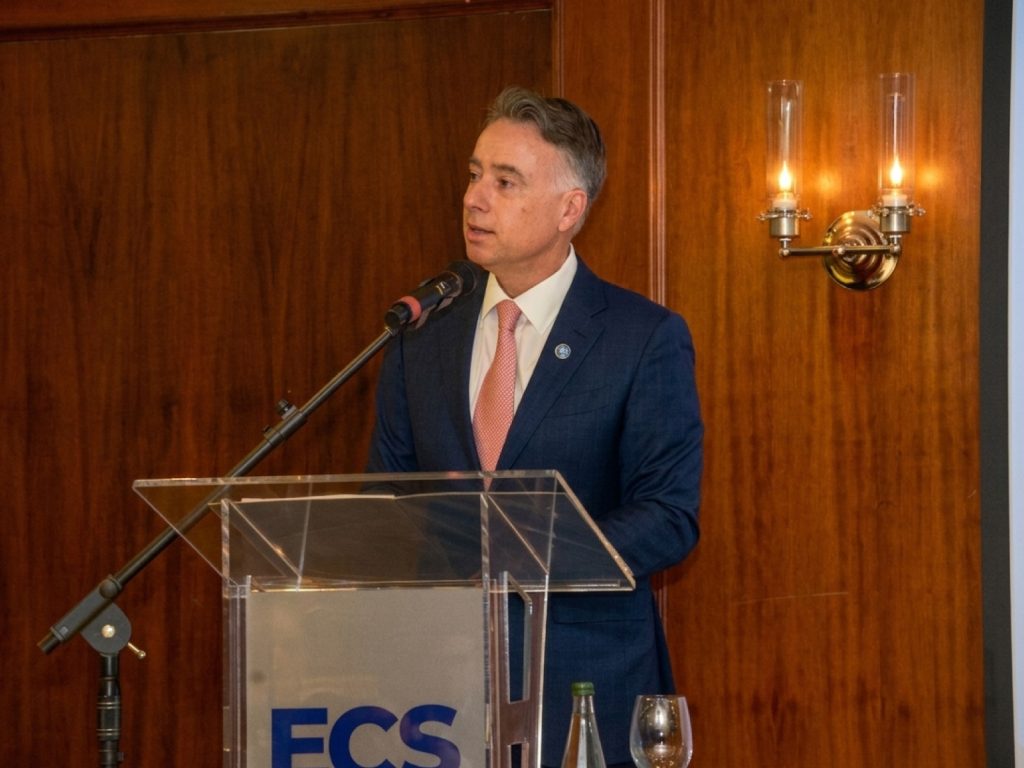 Man in a navy suit and pink tie delivers a speech at a clear lectern with a microphone in a wood-paneled room.