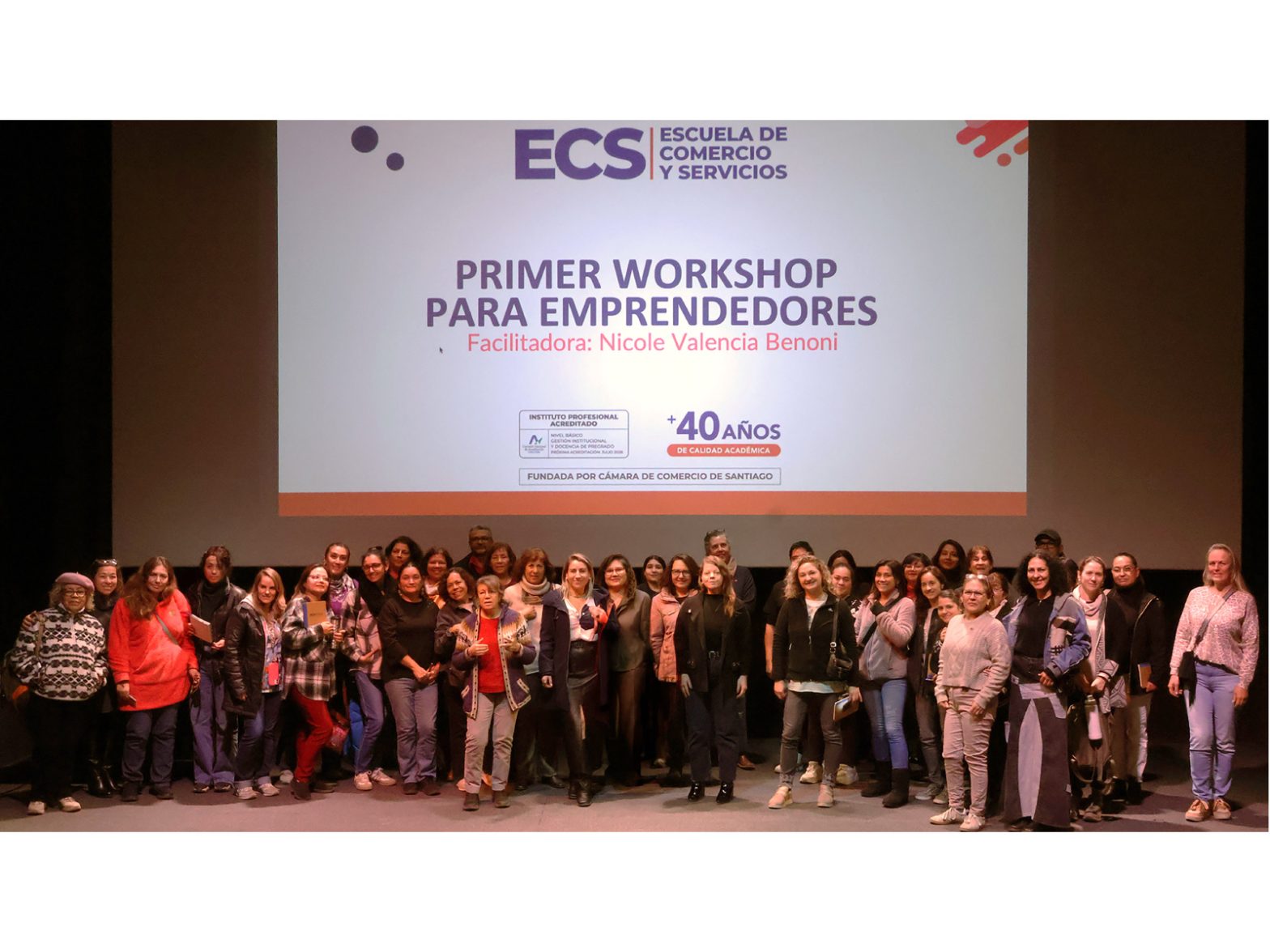 Group of people posing on a stage in front of a large screen showing a workshop title: 'PRIMER WORKSHOP PARA EMPRENDEDORES' and ECS logo.