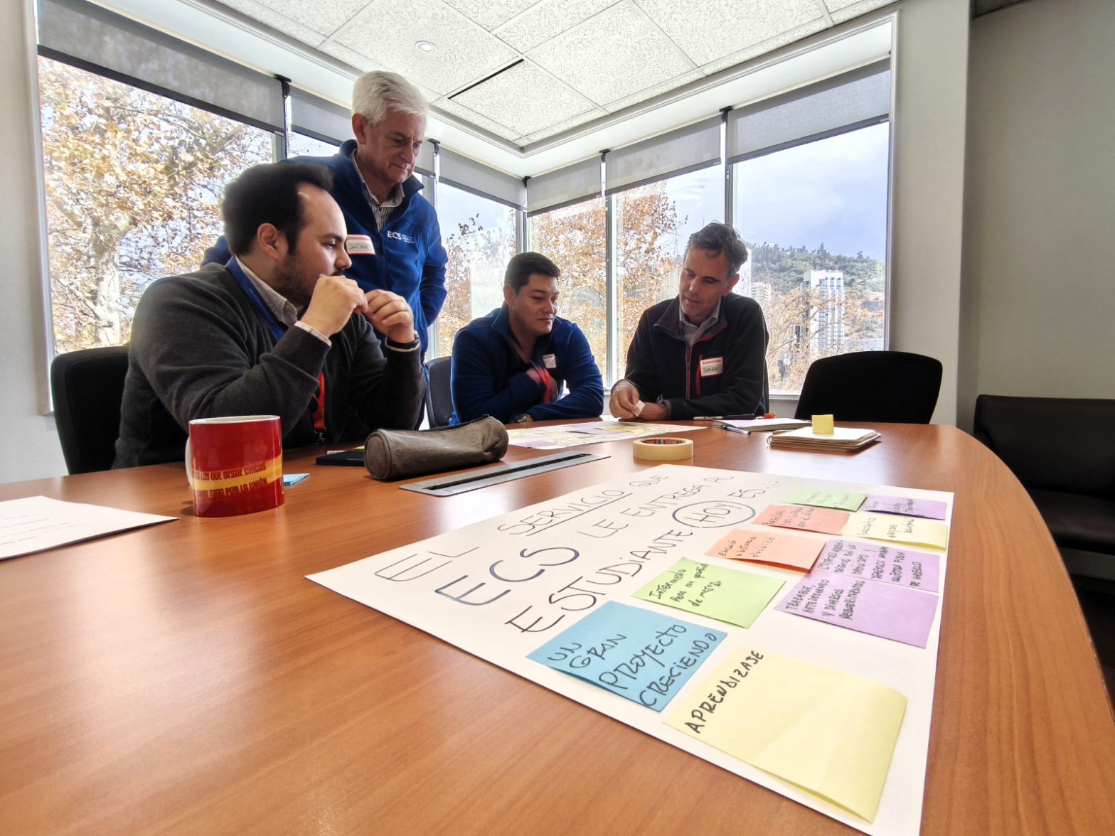 Four men in a meeting room discuss a large poster covered with colorful sticky notes on a wooden table near a windowed wall.