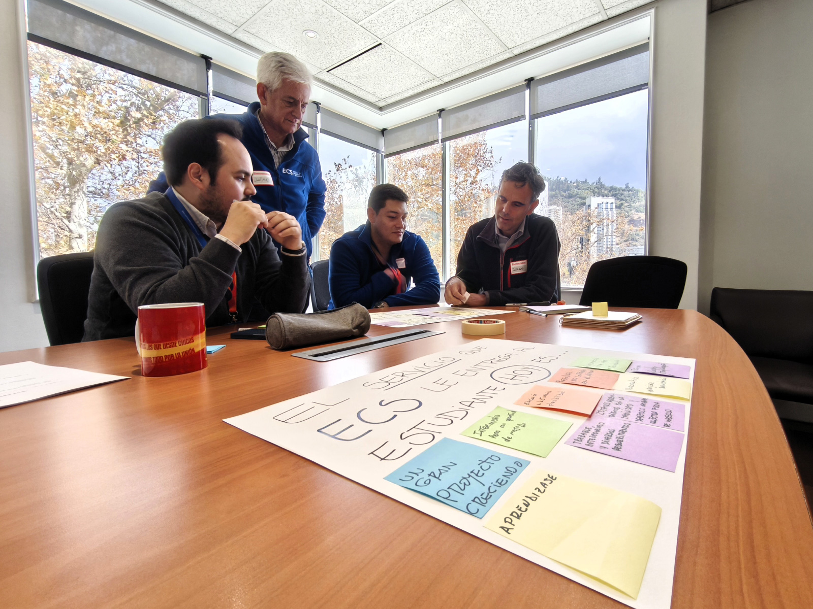 Four men in a meeting room discuss a large poster covered with colorful sticky notes on a wooden table near a windowed wall.