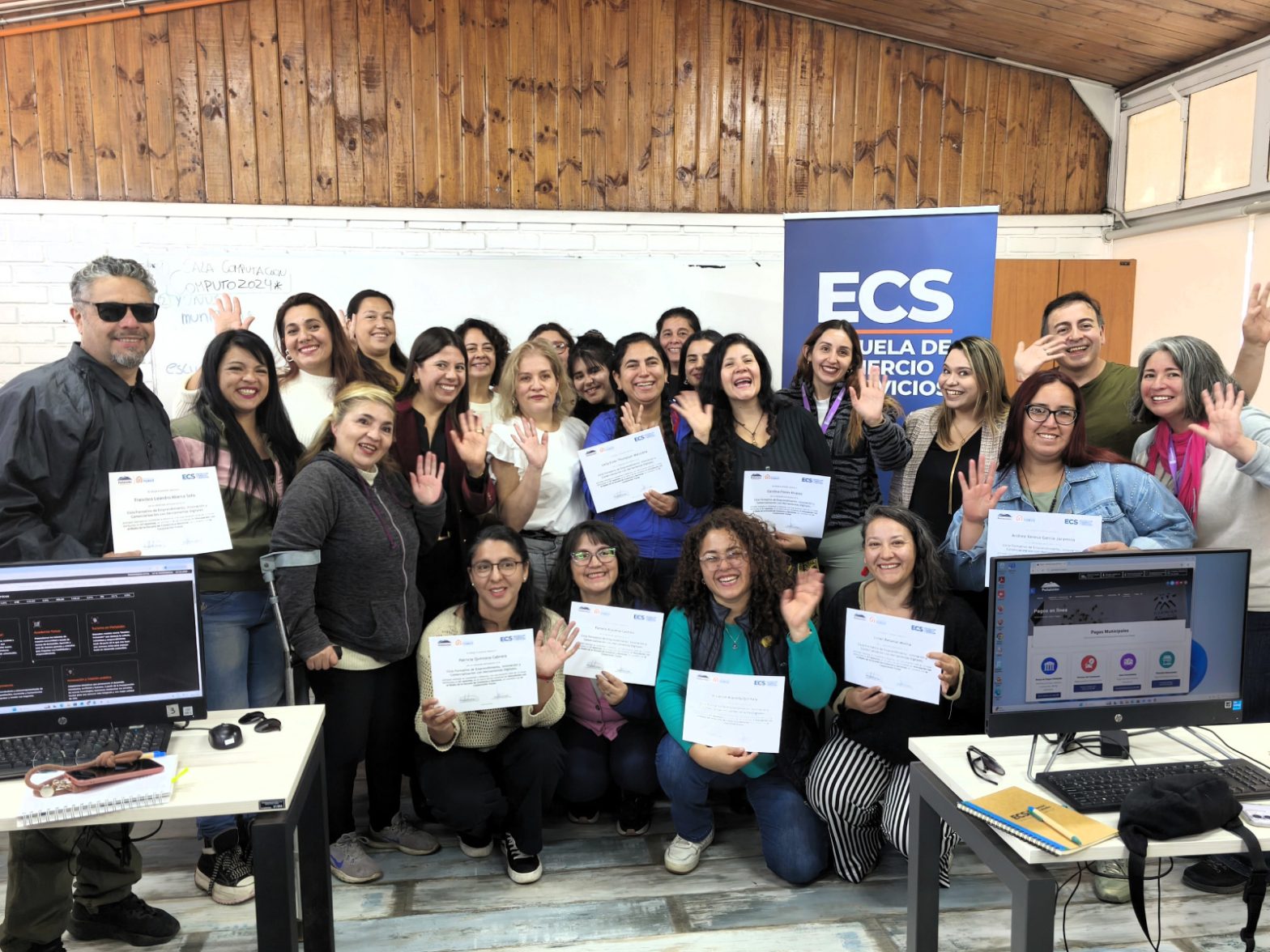 Group of smiling adults indoors, holding certificates and waving for a group photo at an ECS training event with a blue ECS banner visible in the background.