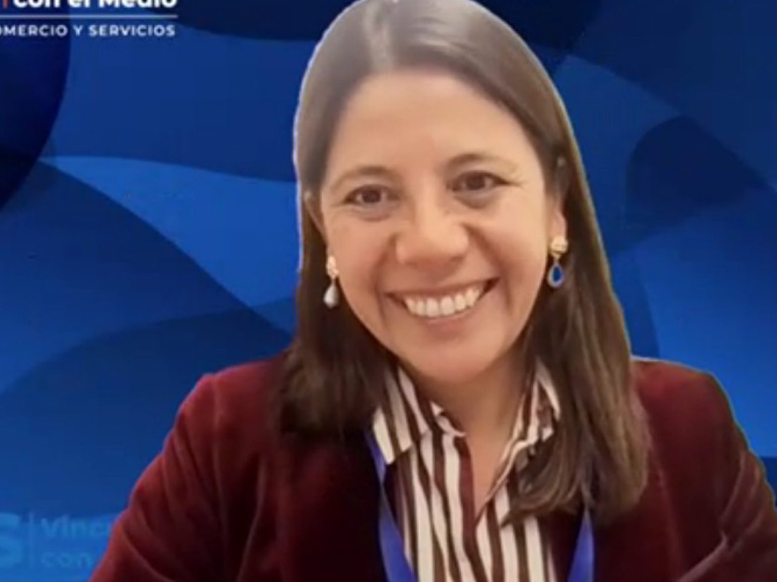 Smiling woman in a burgundy blazer with a striped shirt, against a blue studio backdrop.
