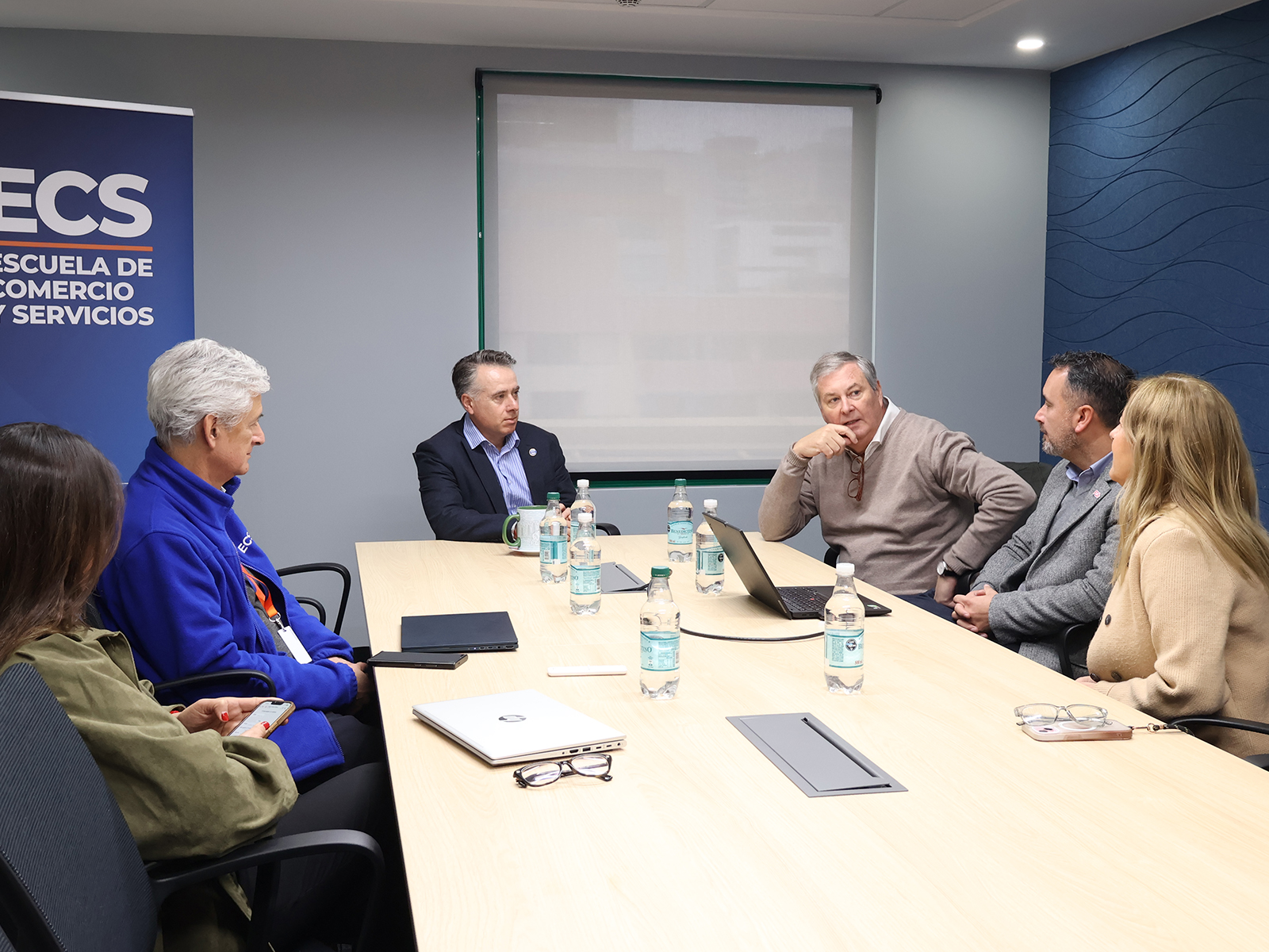 Six people sit around a long conference table in a meeting room, discussing with laptops and water bottles on the table, banner on the left.