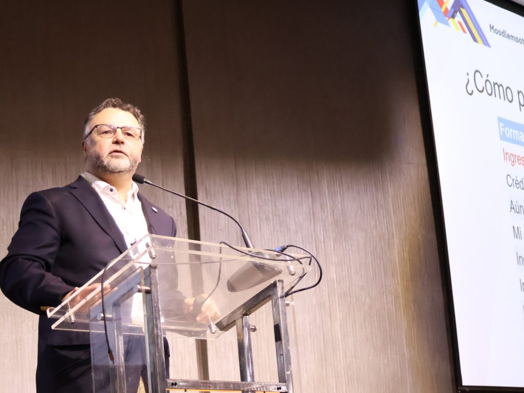 Man in a dark blazer speaking at a clear acrylic podium with a microphone, next to a projected presentation screen.