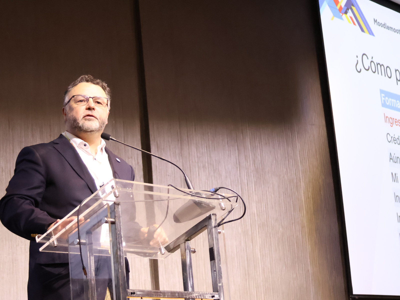 Man in a dark blazer speaking at a clear acrylic podium with a microphone, next to a projected presentation screen.