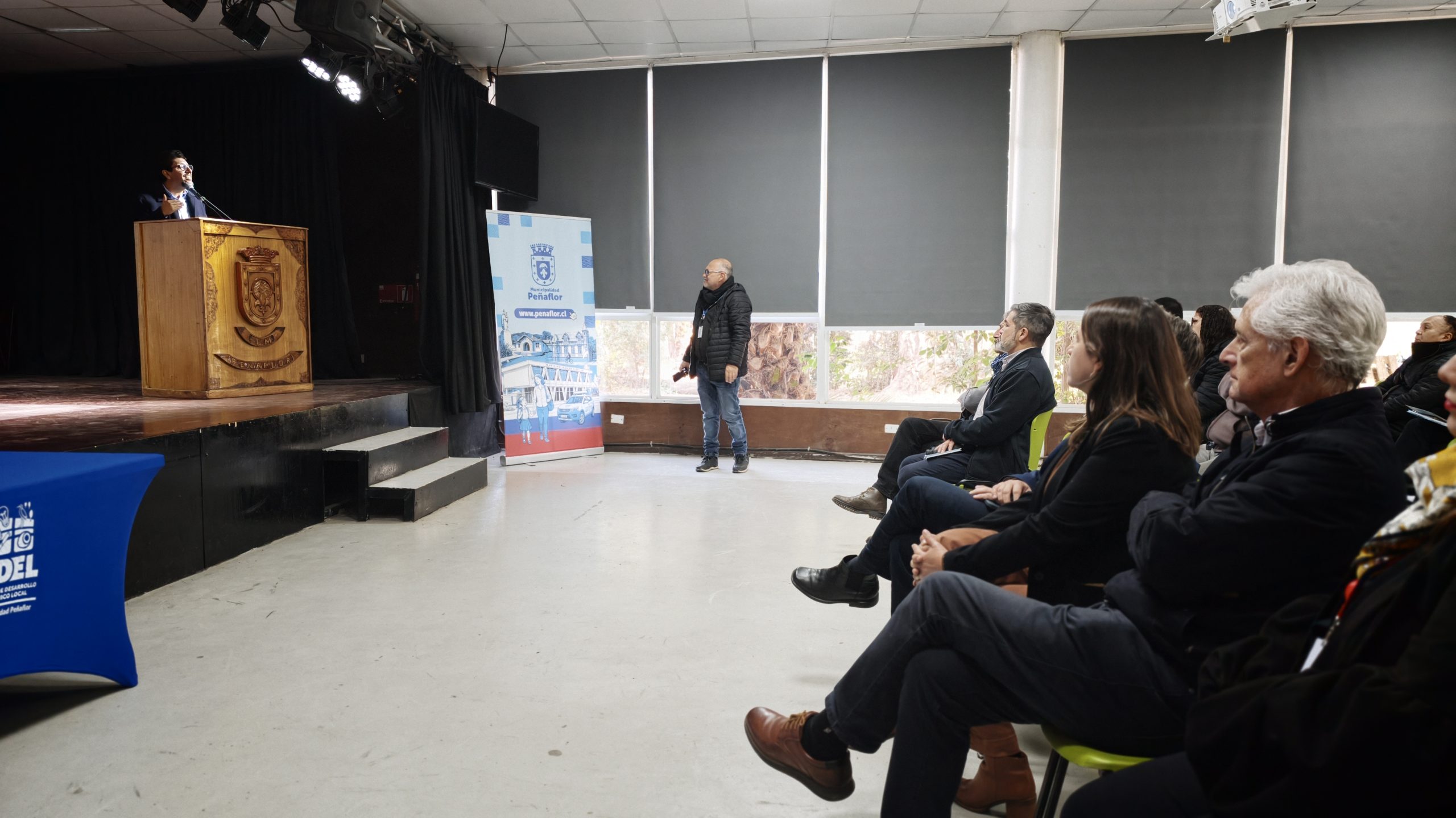 Man at a wooden lectern giving a talk to an audience in a small hall, crest on the front of the podium.