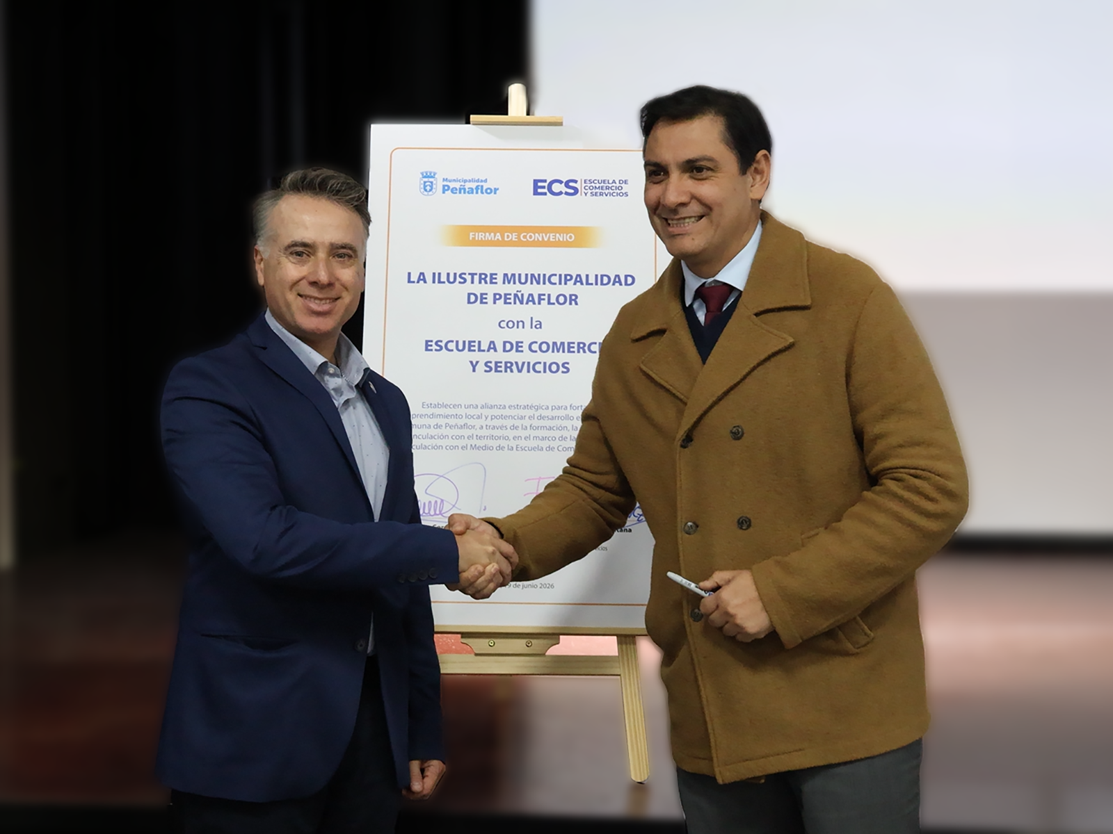 Two men in business attire shake hands beside a sign announcing a agreement between La Ilustre Municipalidad de Peñaflor and the Escuela de Comercio y Servicios.