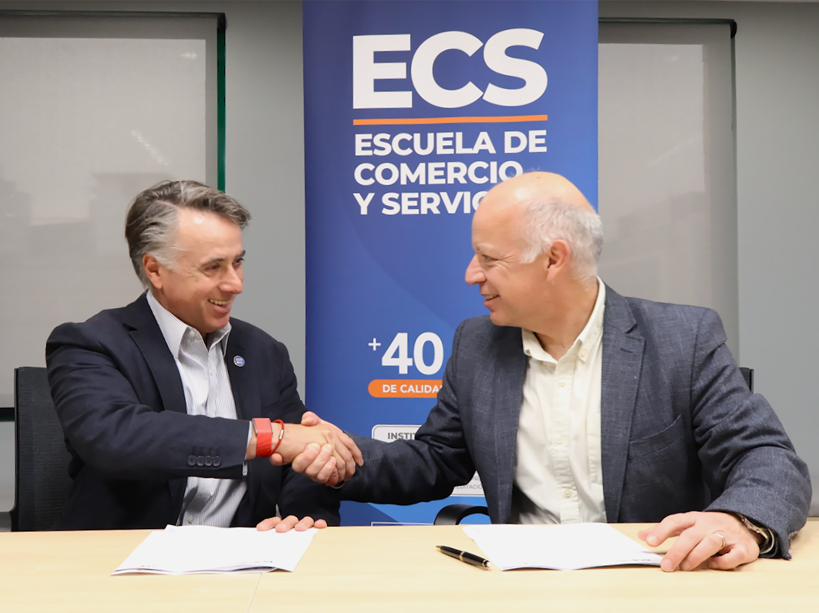 Two men in business attire shake hands across a table in front of an ECS banner.