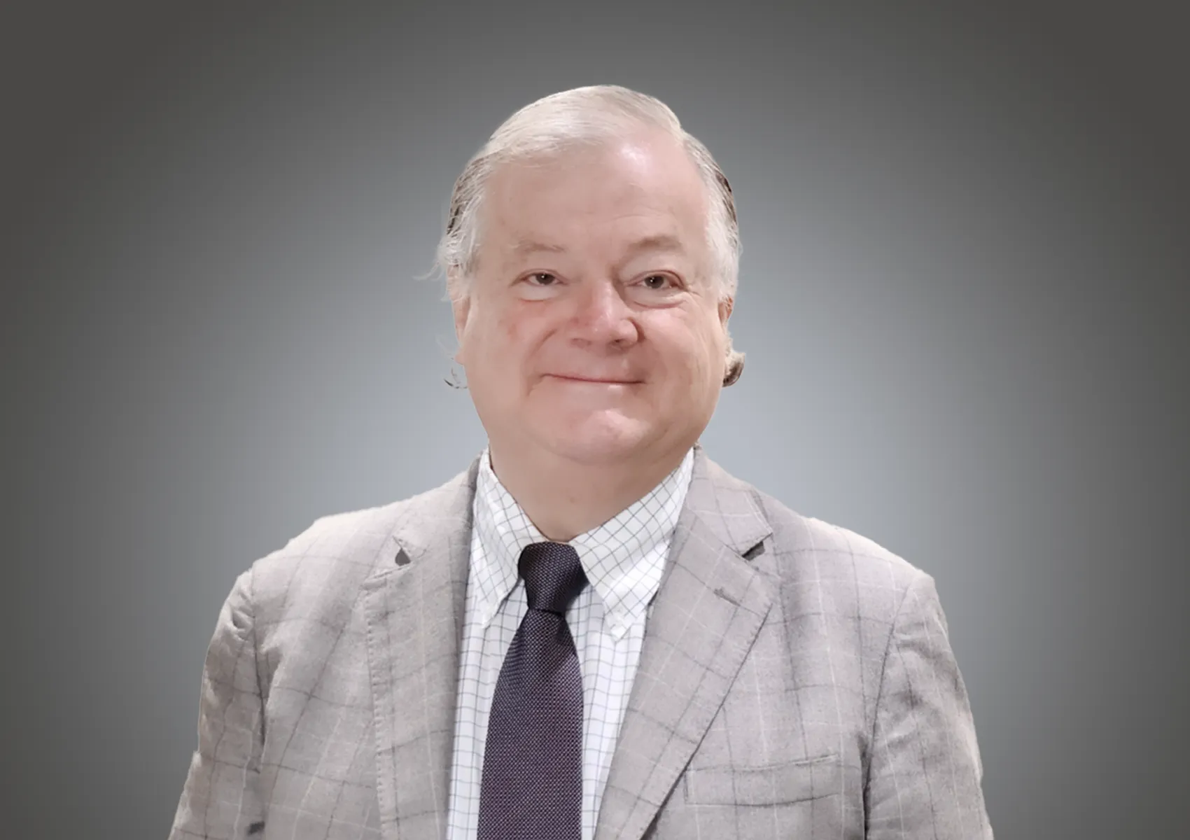 Smiling older man in a light gray plaid suit, white shirt, and dark tie, posed against a gray gradient background.