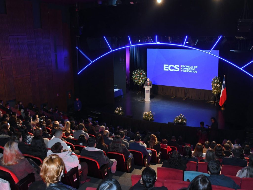 Speaker stands at a podium on a blue-lit stage with a large screen that reads ECS Escuela de Comercio y Servicios; a Chilean flag and flower arrangements flank the stage while an audience sits in a dark theater.