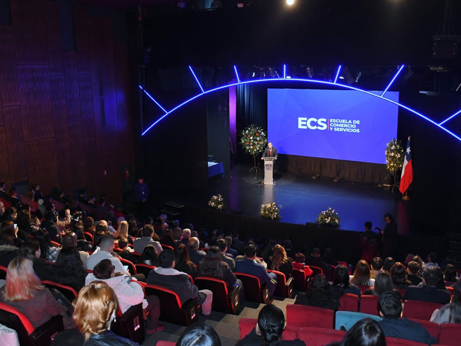 Speaker stands at a podium on a blue-lit stage with a large screen that reads ECS Escuela de Comercio y Servicios; a Chilean flag and flower arrangements flank the stage while an audience sits in a dark theater.