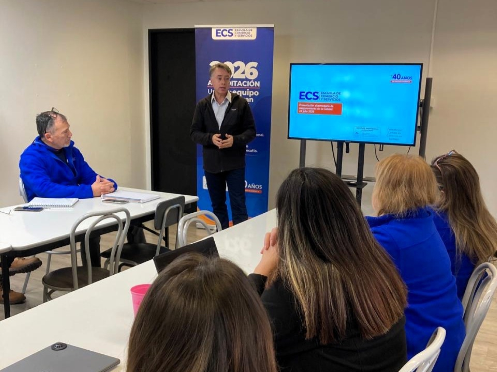 Man wearing a dark jacket presenting to a classroom audience, with a blue ECS banner and a large monitor displaying a blue slide in the background.