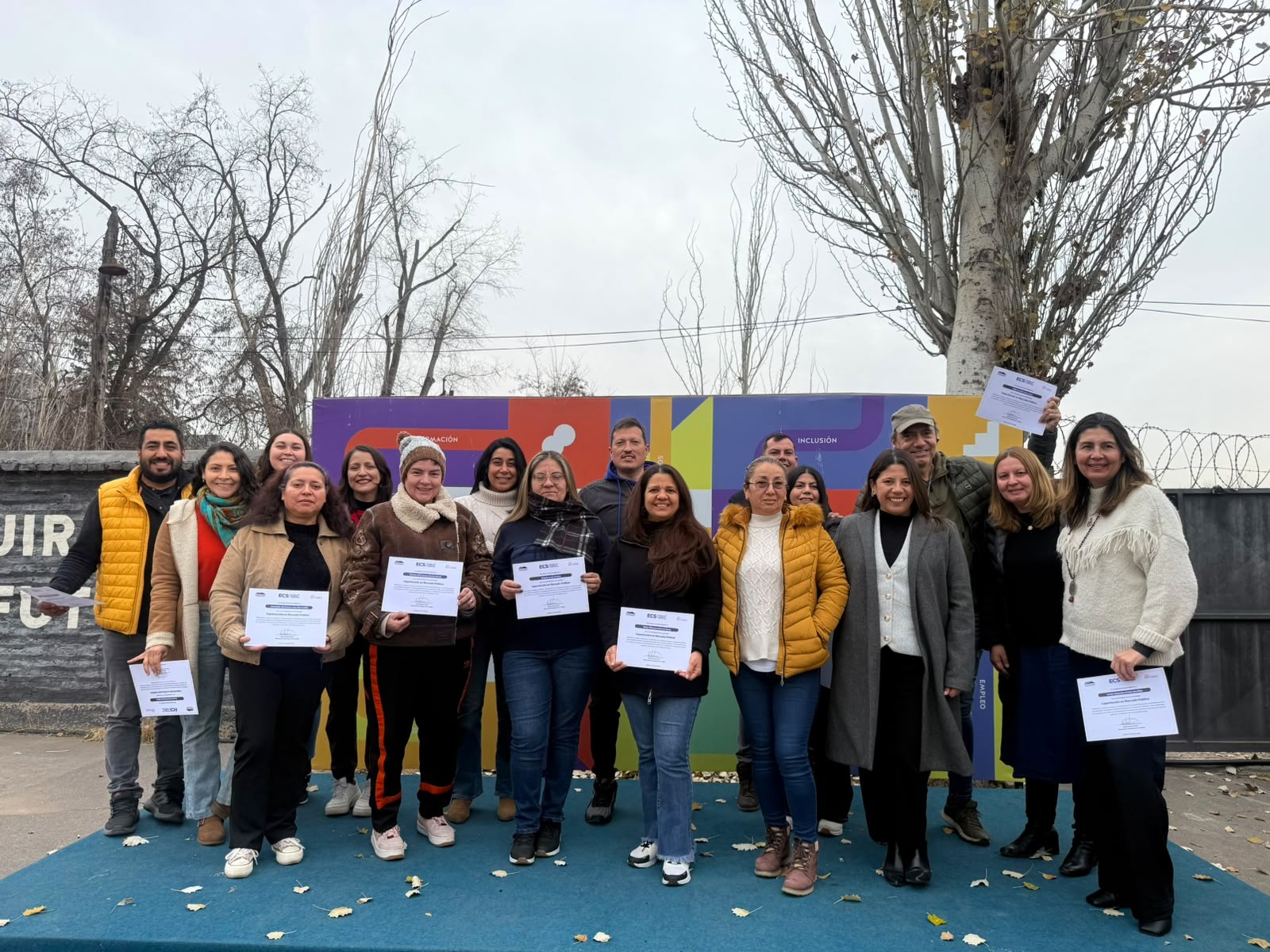 Group of adults posing on a blue outdoor stage, holding certificates during a ceremony in front of a colorful backdrop.