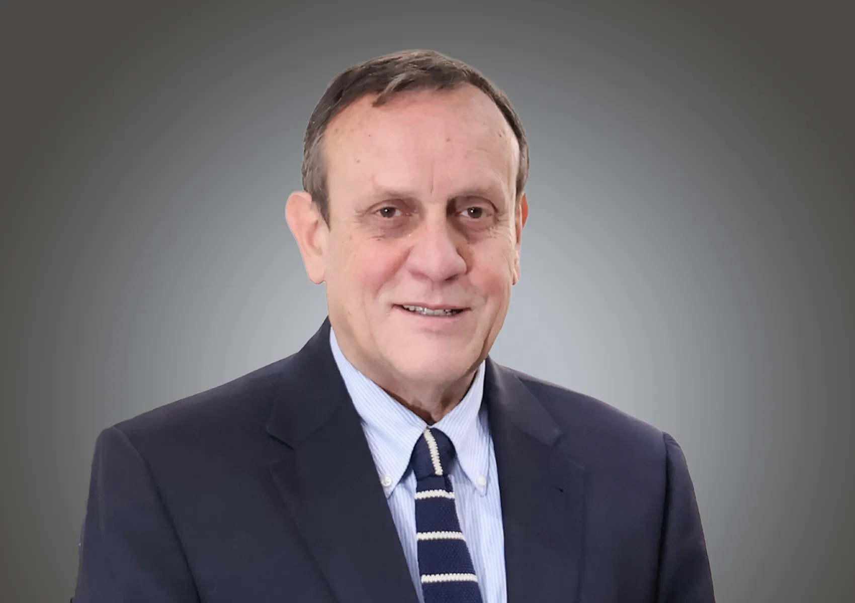 Portrait of a middle-aged man in a dark suit and striped tie, smiling at the camera against a neutral gray background.