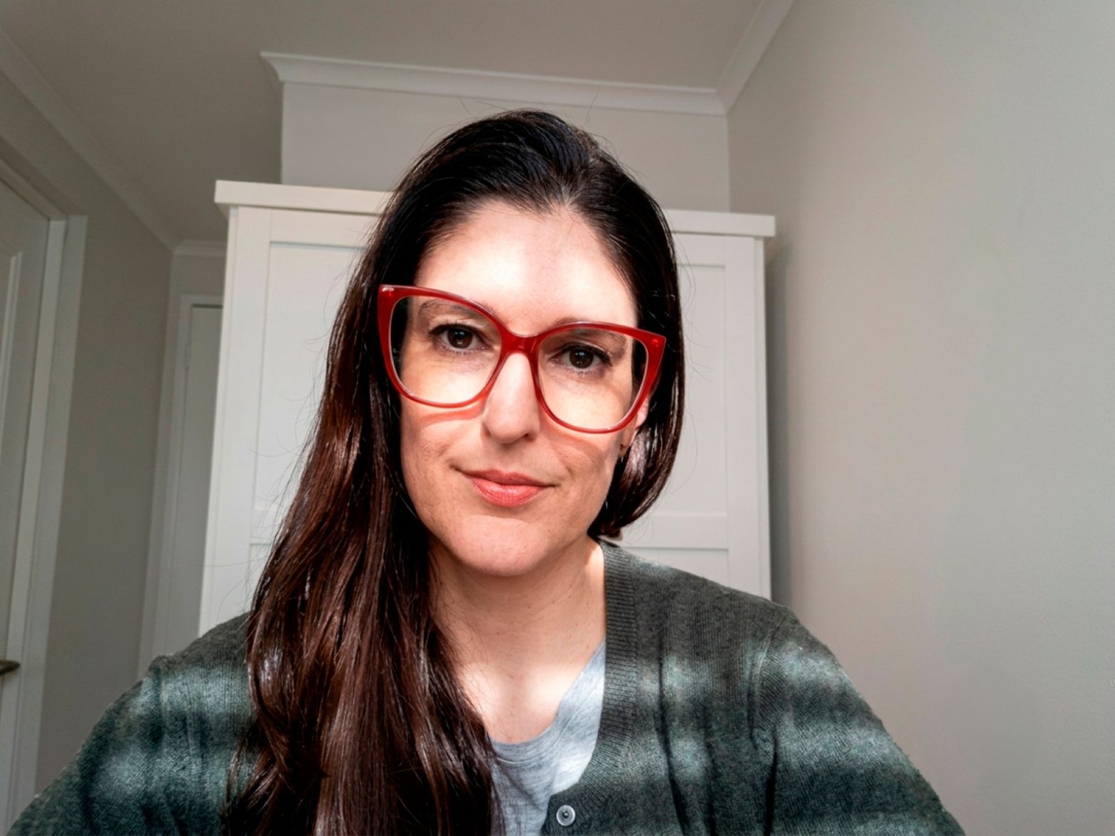 Woman with long dark hair and red glasses poses for a selfie indoors, wearing a green cardigan over a gray top.