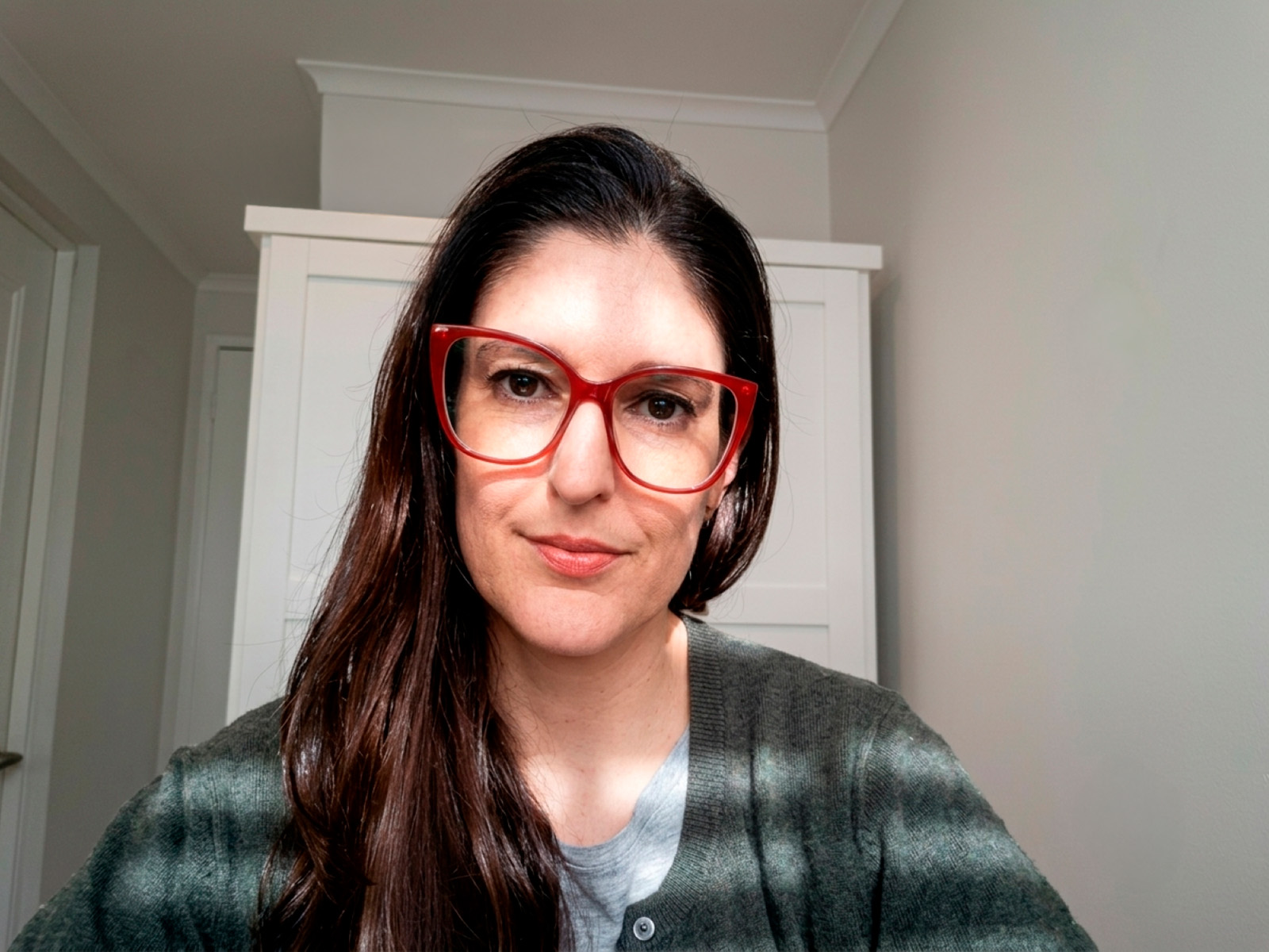 Woman with long dark hair and red glasses poses for a selfie indoors, wearing a green cardigan over a gray top.