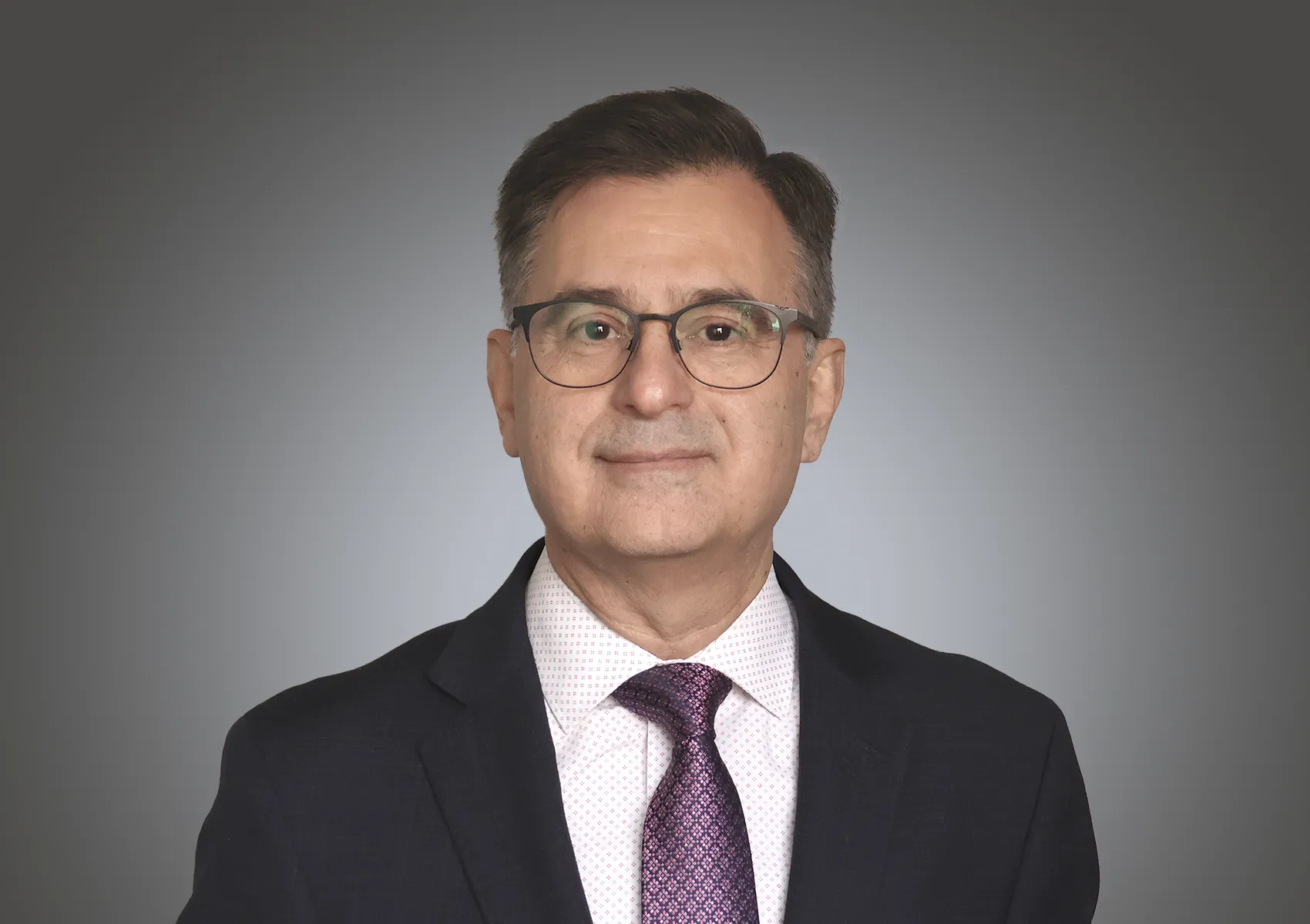 Professional headshot: man in a dark suit, pink patterned tie, glasses, against a gray gradient background.