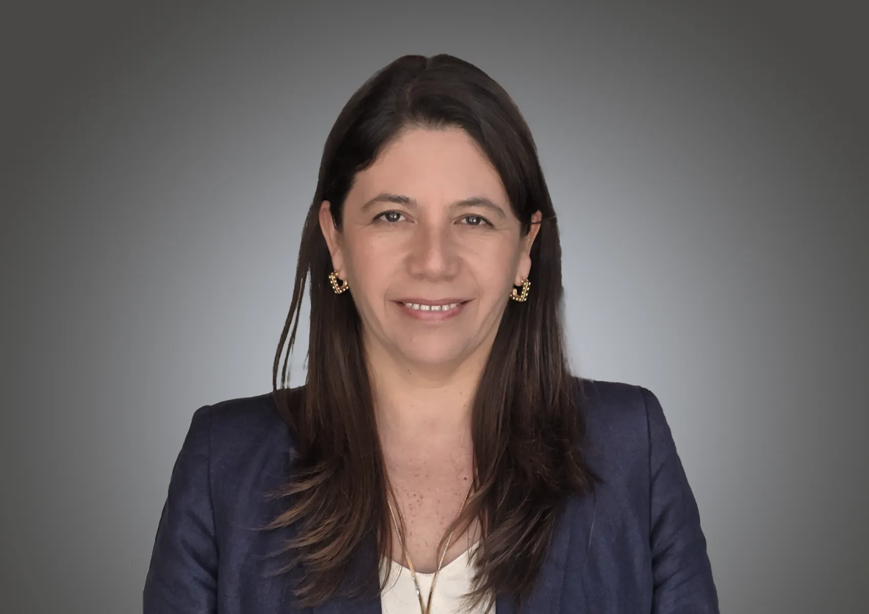 Smiling woman with long dark hair wearing a navy blazer and gold earrings against a gray studio background.