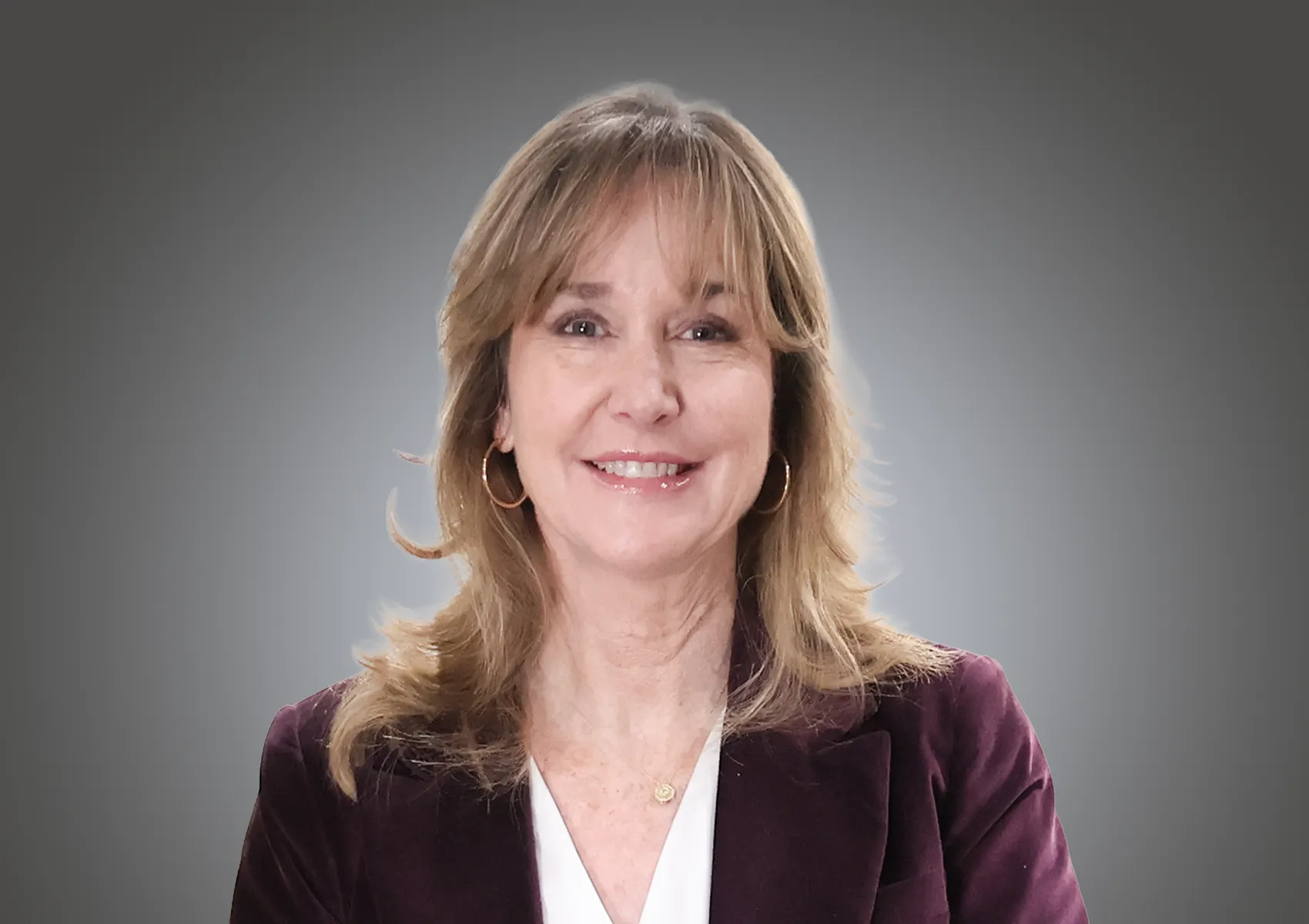 Smiling woman with shoulder-length blond hair in a burgundy blazer and white blouse, wearing hoop earrings, against a gray gradient background.
