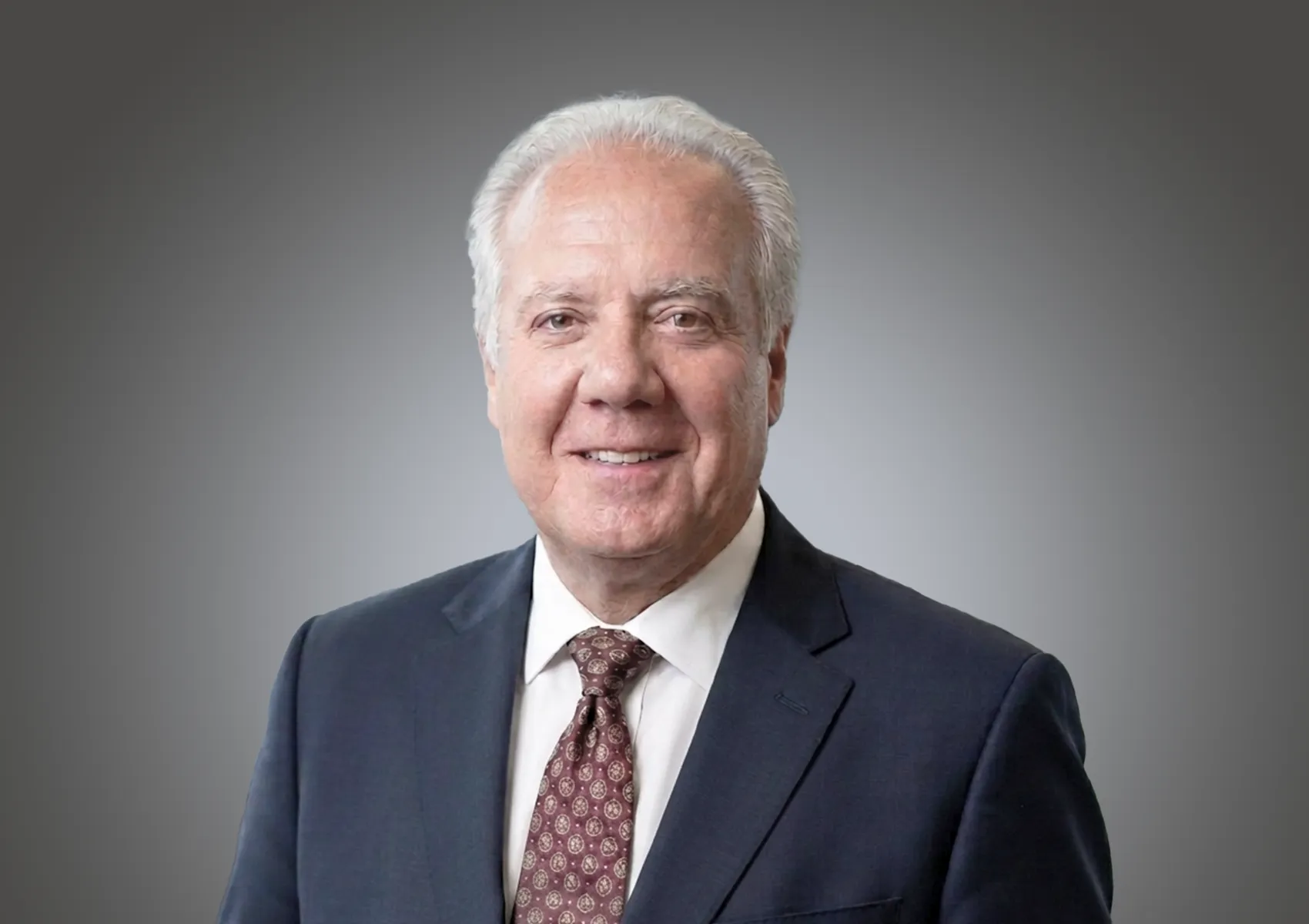 Smiling older man in a navy suit, white shirt, and maroon patterned tie against a gray gradient backdrop (business headshot).