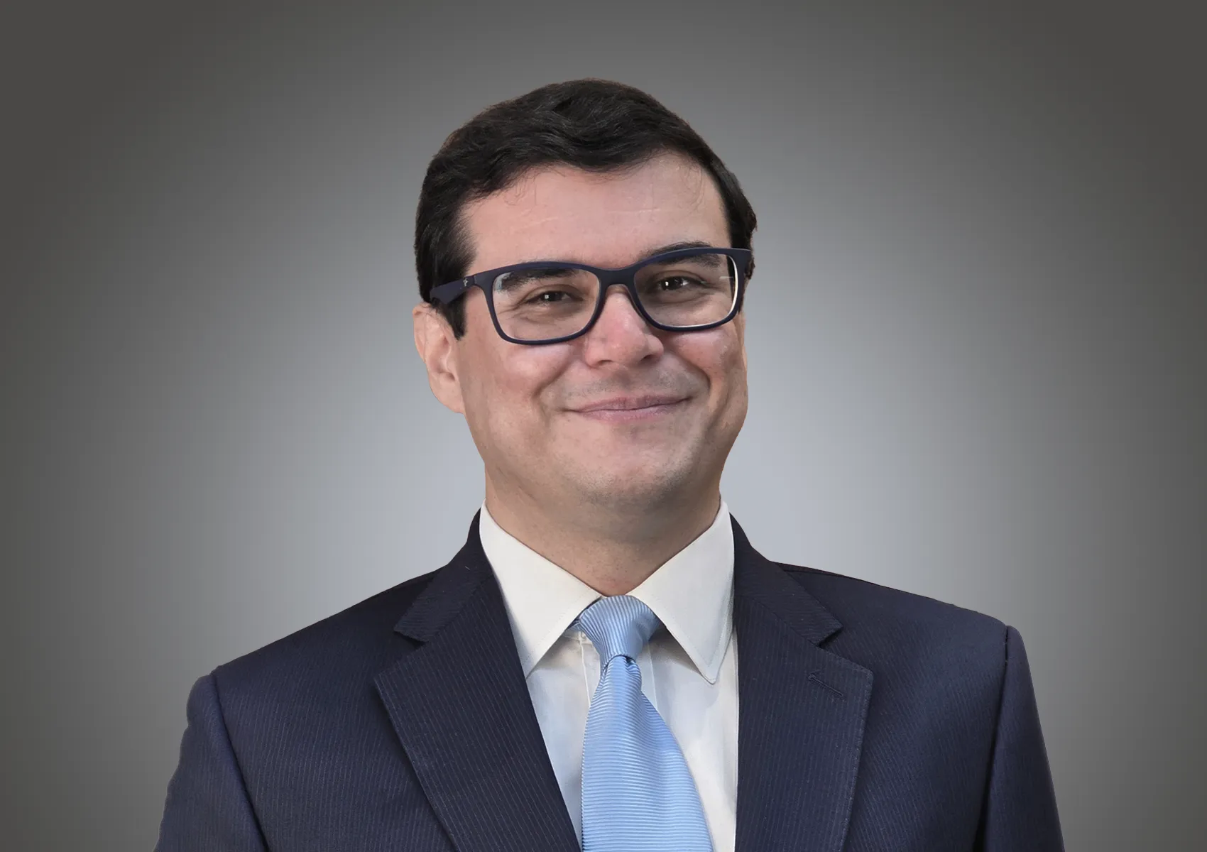 Smiling man in a navy suit, light blue tie, and glasses against a gray gradient background (head-and-shoulders portrait).