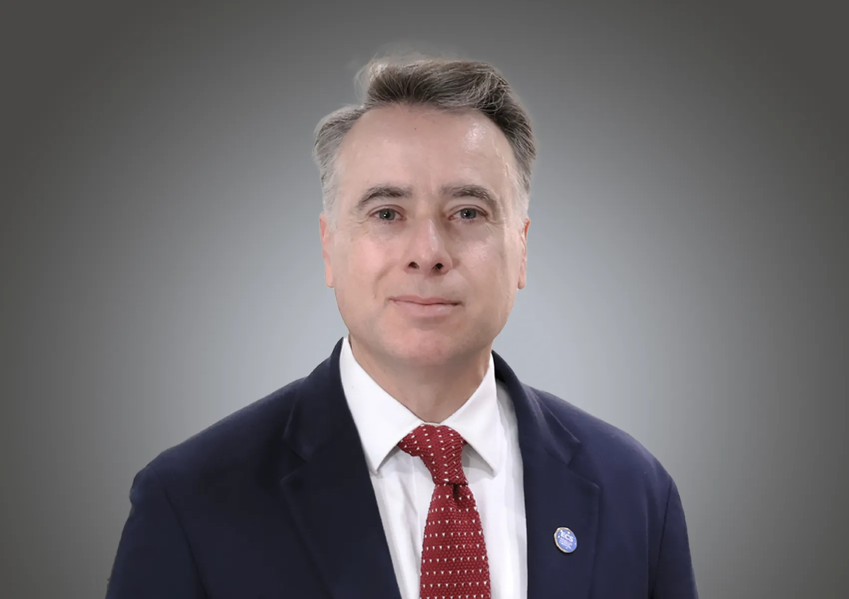 Professional head-and-shoulders portrait of a man in a dark suit and red tie against a gray gradient background.