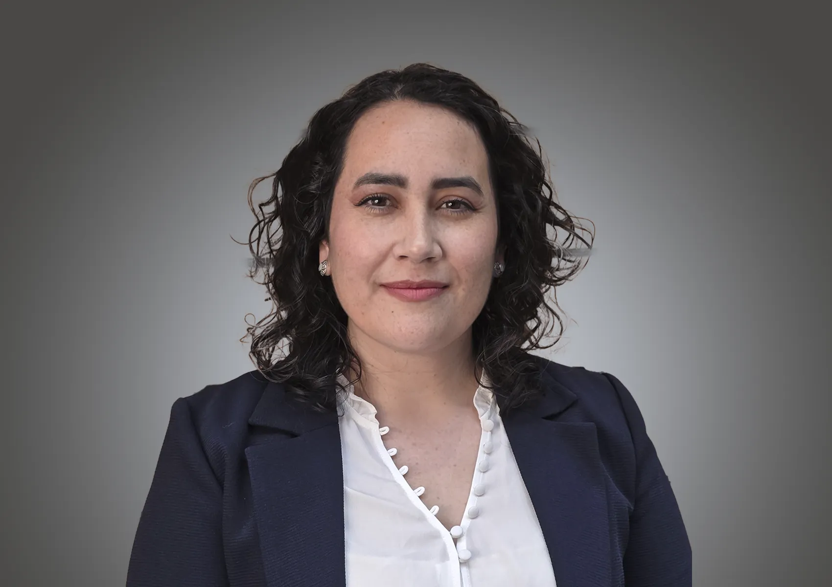 Portrait of a woman with dark curly hair, wearing a navy blazer and white blouse, smiling at the camera against a gray gradient background.