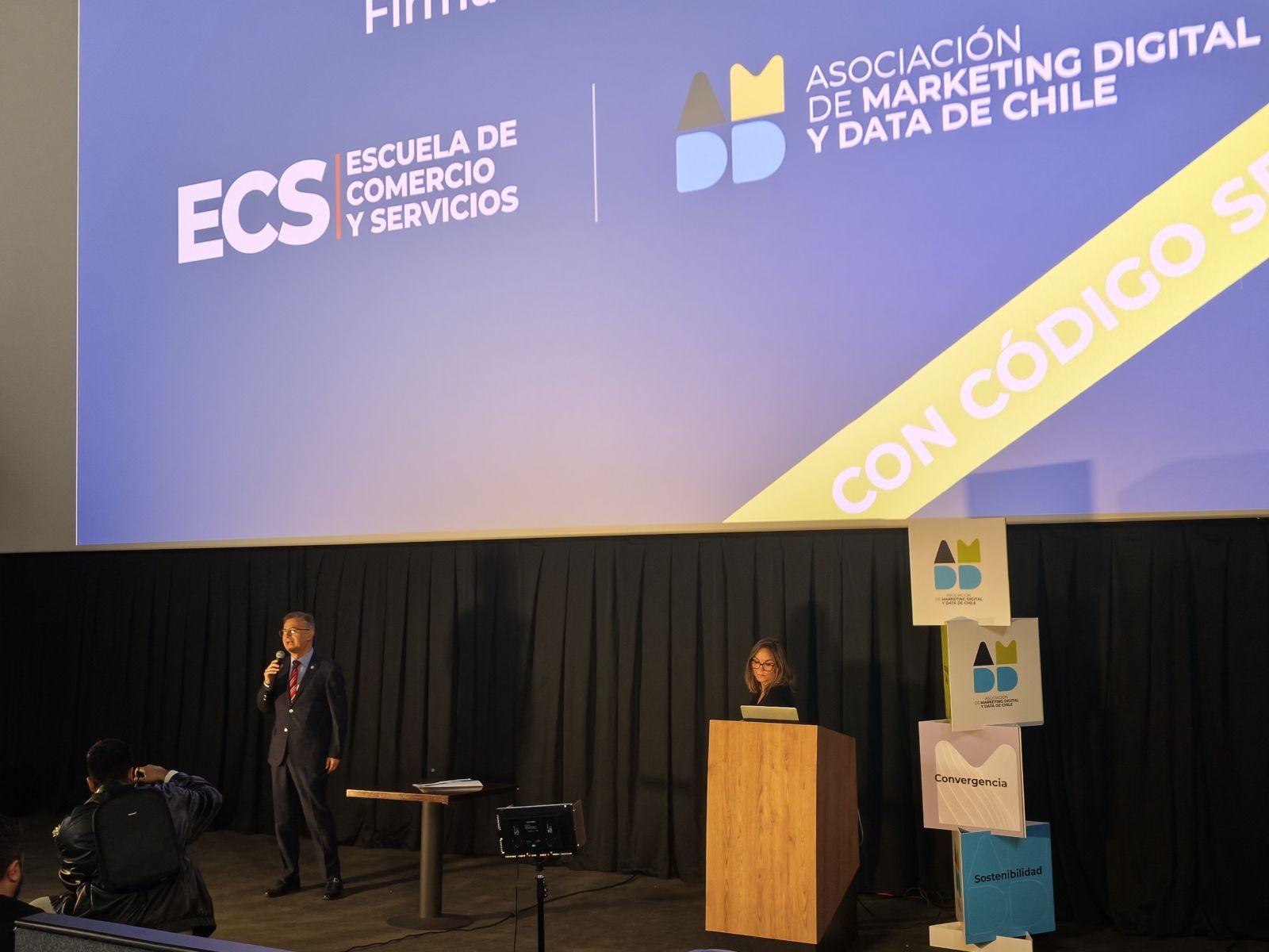 Speaker in a suit addresses the audience on a stage with a large blue banner featuring ECS and Asociación de Marketing Digital y Data de Chile logos behind him, and a woman stands at a podium nearby.