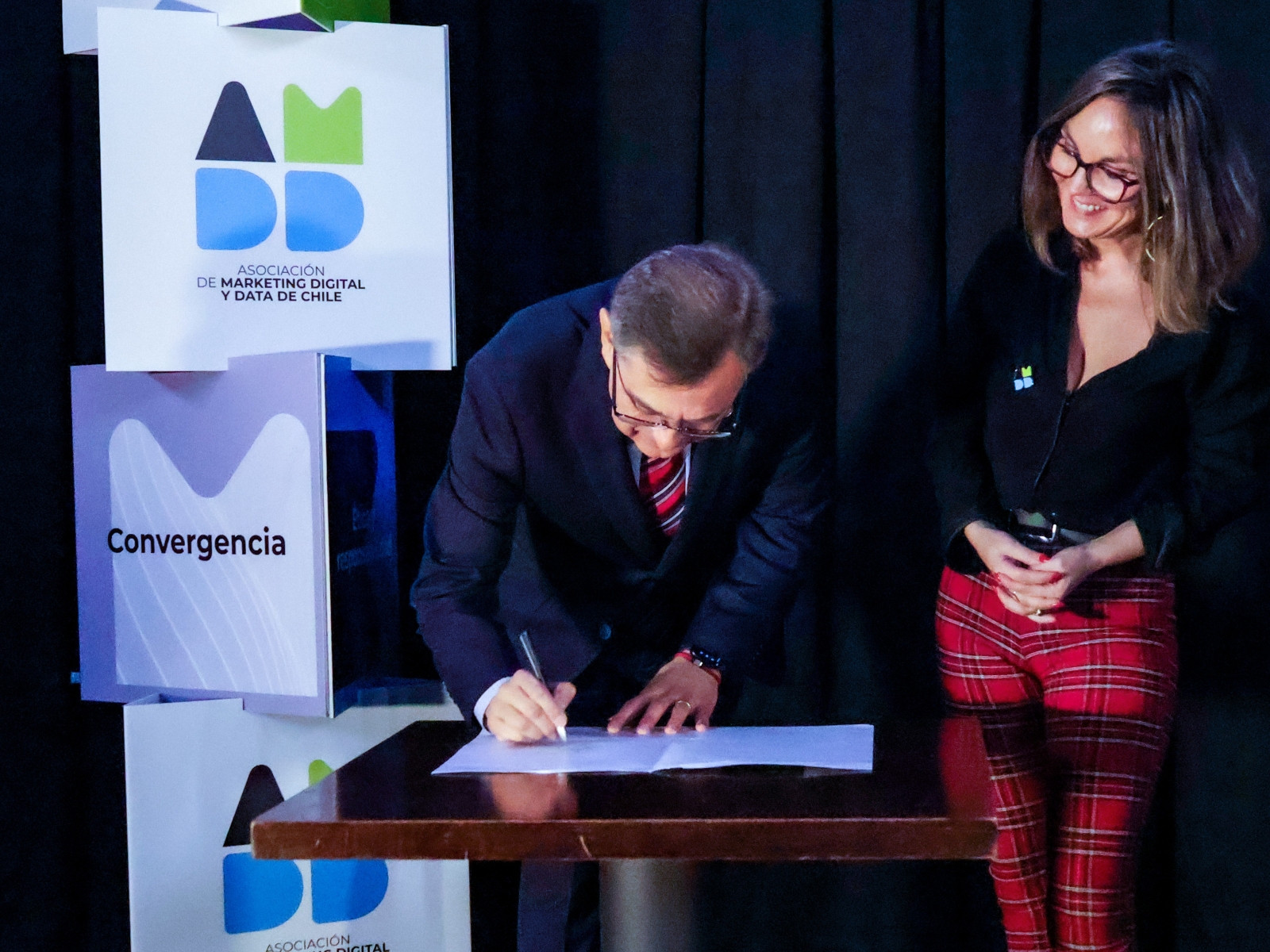 Man in a dark suit signs a document at a wooden podium as a smiling woman in glasses watches nearby, with Convergencia banners in the background.