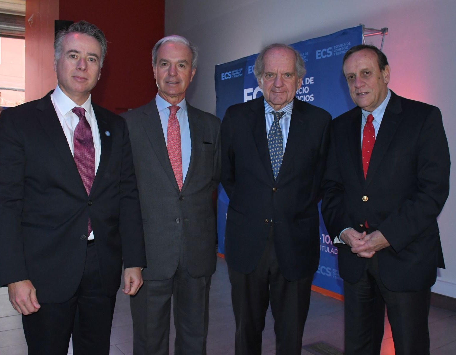 Four men in dark suits posing for a photo at a formal event, with a blue banner in the background.