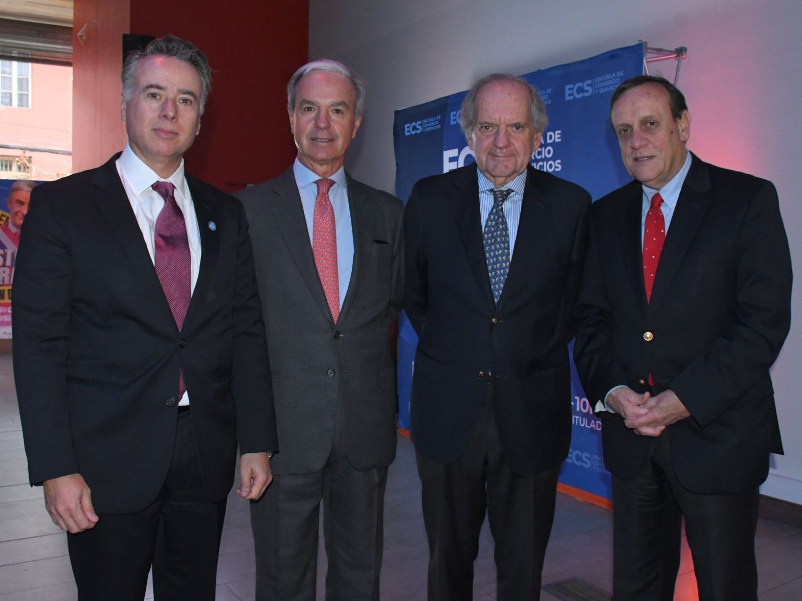 Four men in dark suits posing for a group photo at an event, with a blue ECS banner in the background.