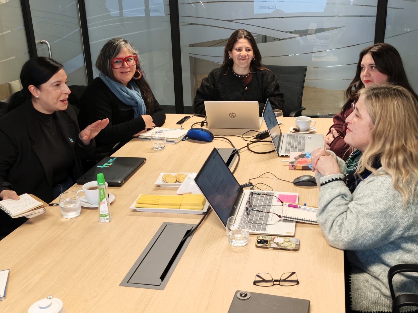 Five women sit around a conference table in a glass-walled meeting room, discussing and working on laptops and documents.