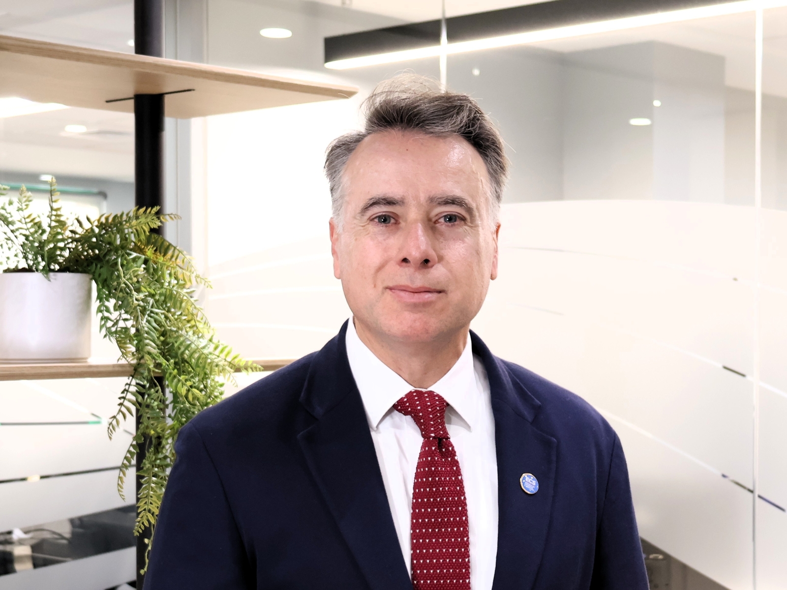 Professional man in a navy suit and red tie stands in a modern office with glass panels and a potted plant nearby.