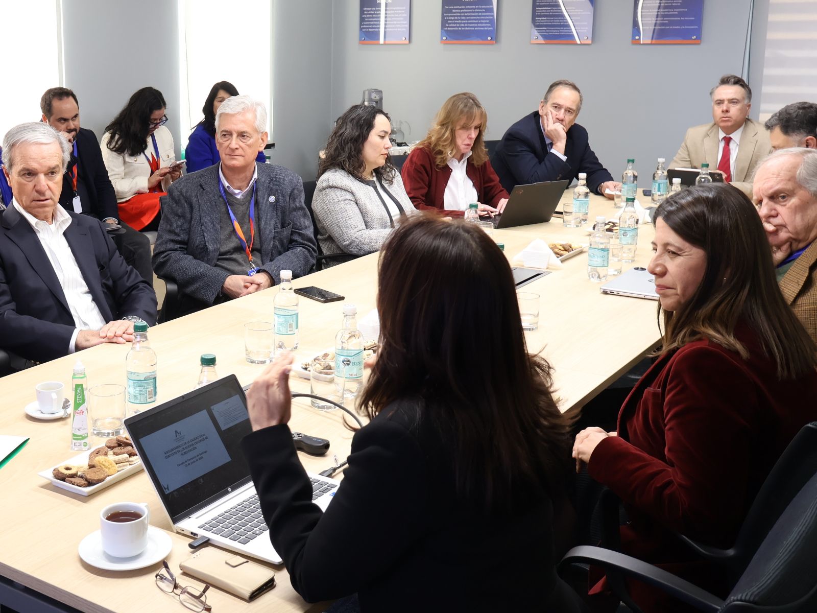 Group of professionals seated around a long conference table, with laptops, water bottles, cups, and snacks during a meeting.