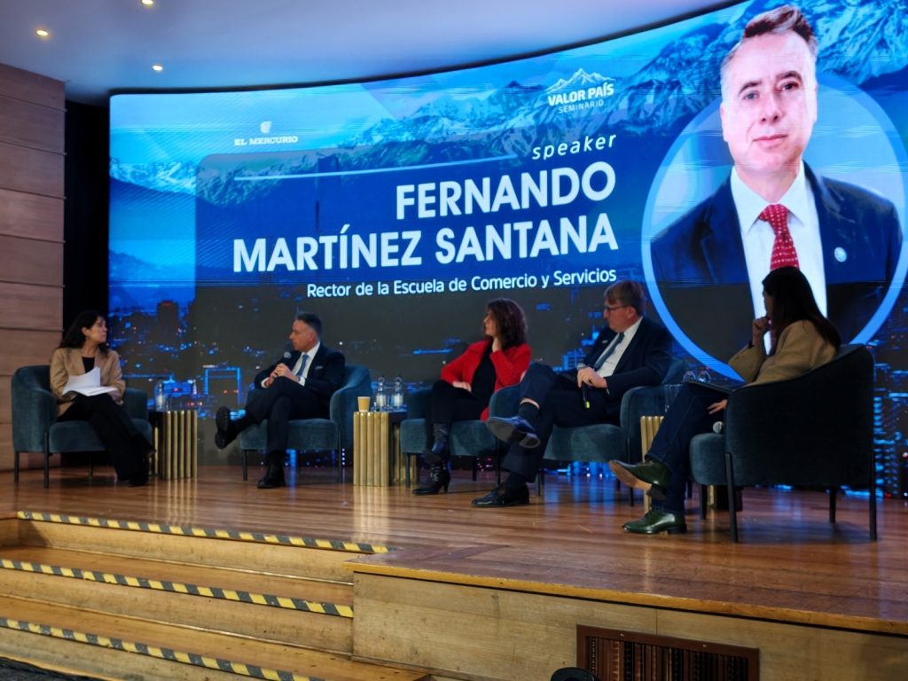 Panel discussion on a stage with five people seated in blue chairs, a large blue backdrop displays the name Fernando Martínez Santana and a speaker portrait.